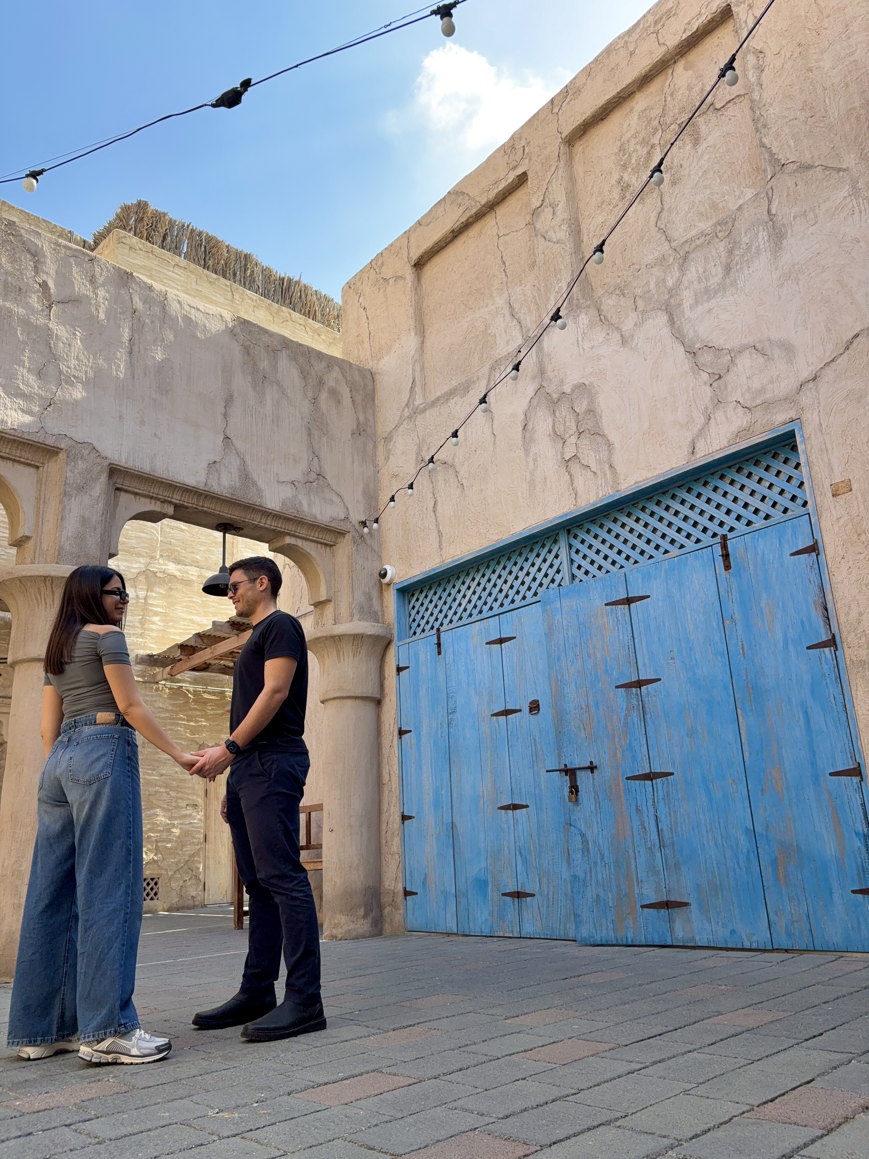 Couple standing facing each other and holding hands in a historic Arabian courtyard with a striking blue door