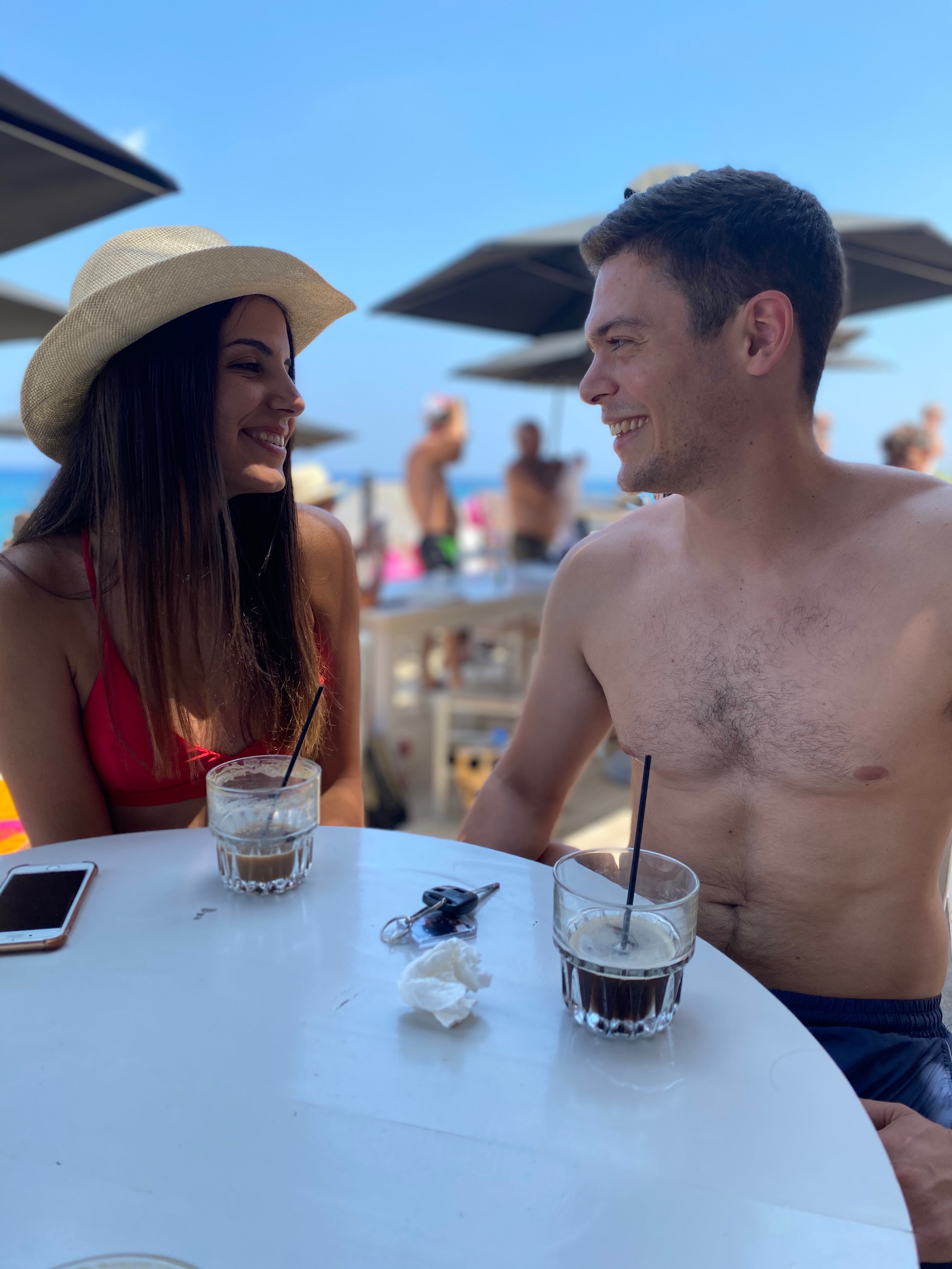 Couple laughing together across a beach café table with iced coffees, golden afternoon light